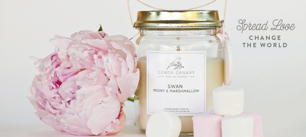 Lemon Canary Swan scented vintage soy candle with 'spread love change the world' text. A pink peony flower is sitting next to the candle with some white and pink marshmallows.